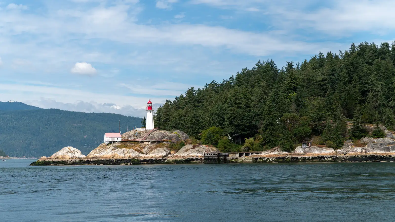 The white Point Atkinson Lighthouse with its red lantern on a rocky point at the edge of Lighthouse Park, with evergreen forest behind it, pale rock along the shore and blue water in front