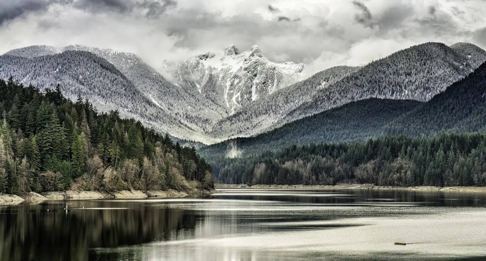 Capilano Lake with forest on both shores and the two snow-covered peaks of the Lions under cloud behind it
