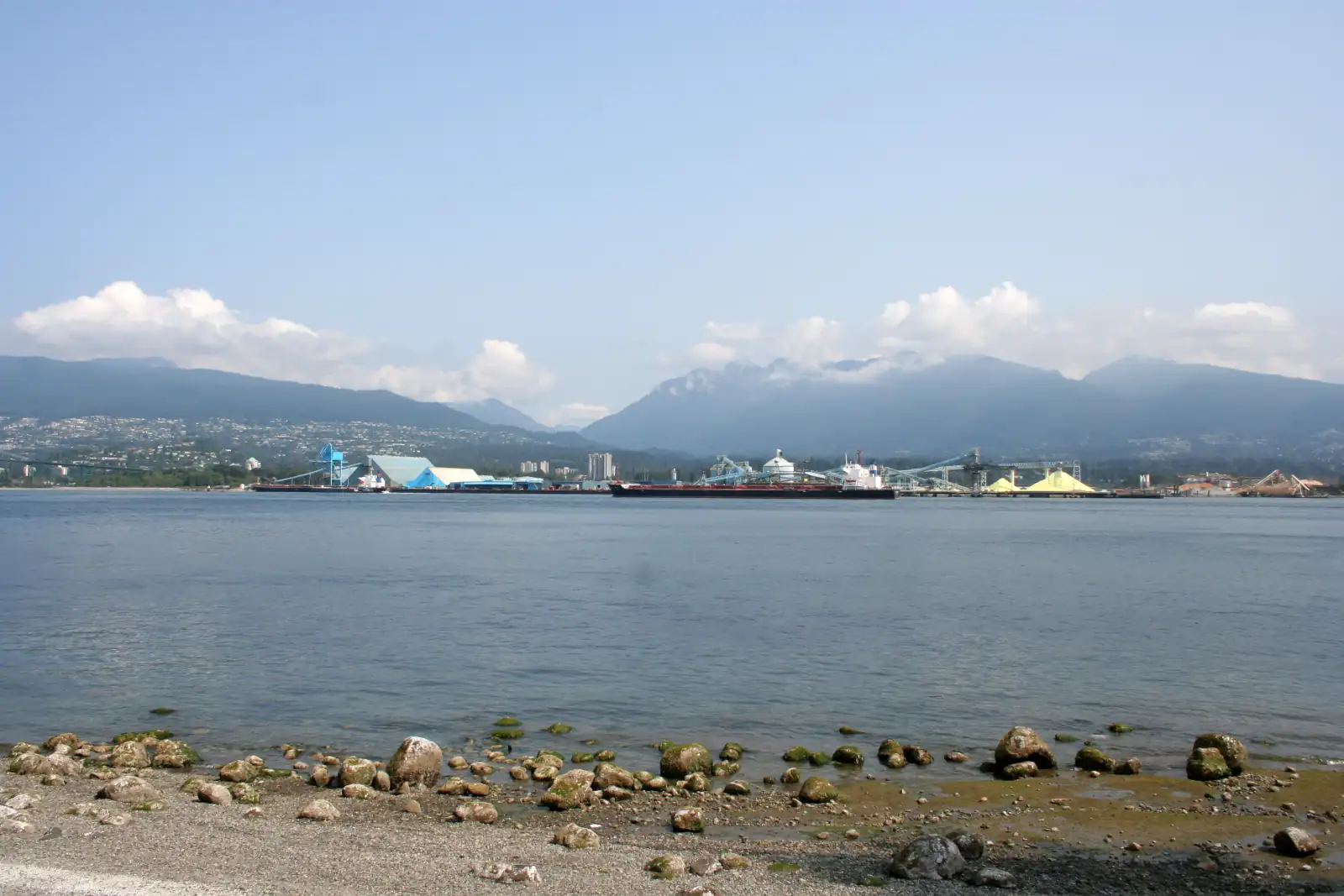 Ships and port terminals on the north shore of Burrard Inlet seen across the water, with forested mountains behind