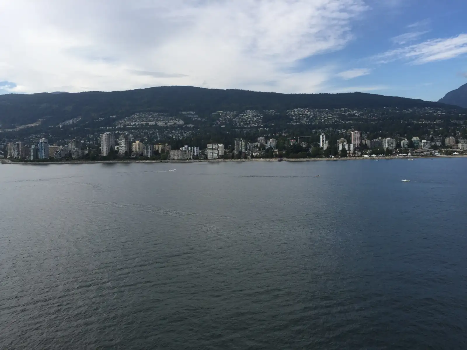 The West Vancouver shore seen from a ship on Burrard Inlet: apartment towers along the water, houses on the tree-covered slope behind them and a forested ridge above under a partly cloudy sky