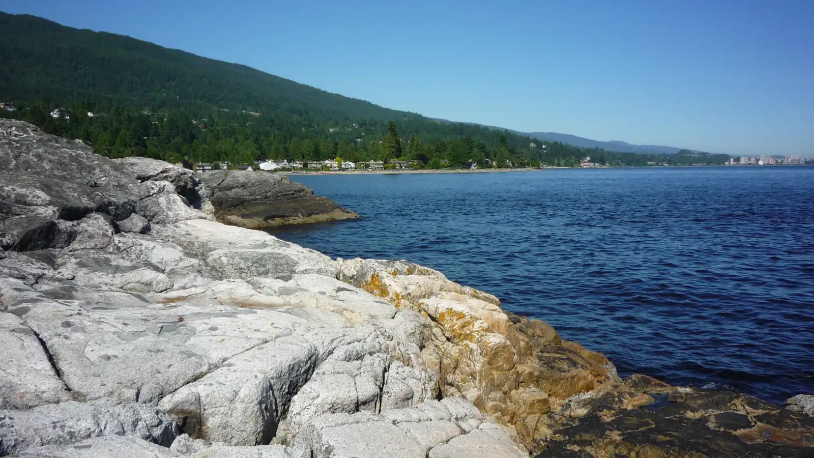 Pale rock on the West Vancouver shore near Caulfeild Park, with blue water, a row of houses behind a beach and a forested hillside rising above them